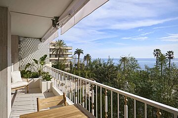 A bright balcony with a white railing overlooks the blue ocean, tropical palm trees, and lush greenery under a clear sky.