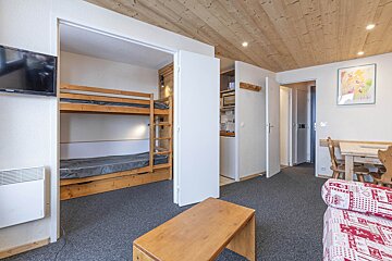A studio apartment with bunk beds, a compact kitchenette, a dining table, and a patterned sofa. Features a wood ceiling and grey carpet.