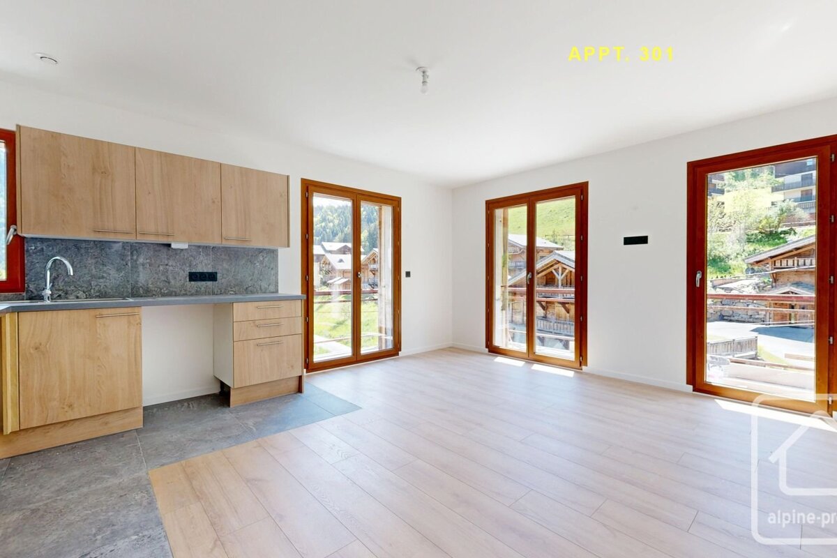 Bright, modern apartment interior with light wood kitchenette, white walls, and large windows showcasing scenic mountain chalet views.