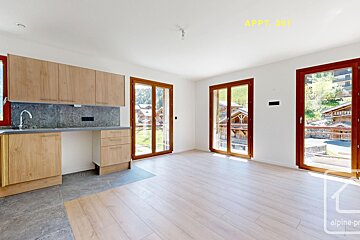 Bright, modern apartment interior with light wood kitchenette, white walls, and large windows showcasing scenic mountain chalet views.