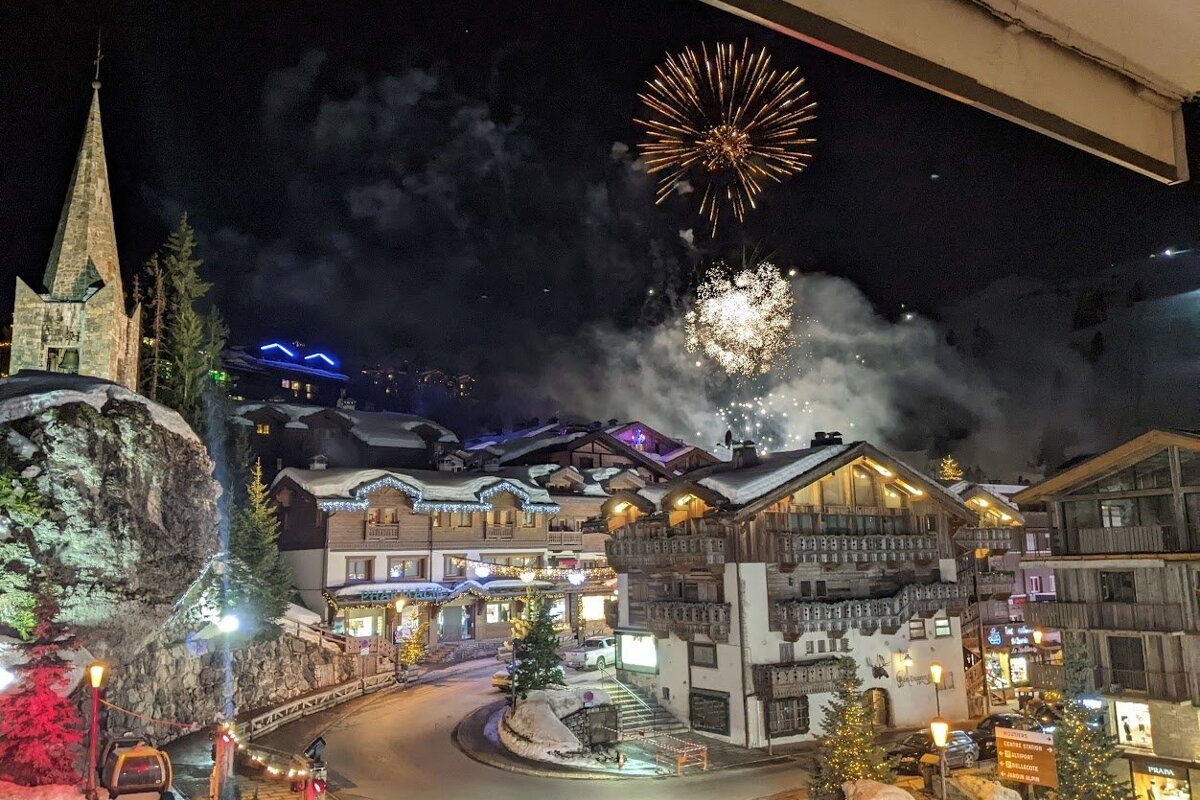 Nighttime fireworks illuminate a picturesque, snow-covered alpine village with a prominent church steeple and festive traditional buildings.