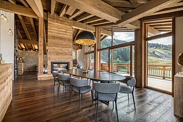 Chic rustic dining room with wooden beams, modern table, fireplace, and floor-to-ceiling windows offering expansive mountain views.