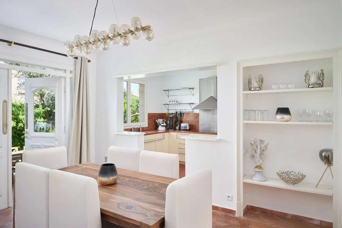 Bright dining area with wooden table, white chairs, and modern chandelier, connected to a kitchen via a pass-through and opening to a lush patio.