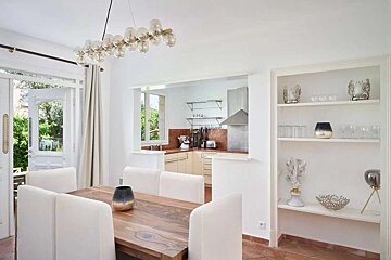 Bright dining area with wooden table, white chairs, and modern chandelier, connected to a kitchen via a pass-through and opening to a lush patio.