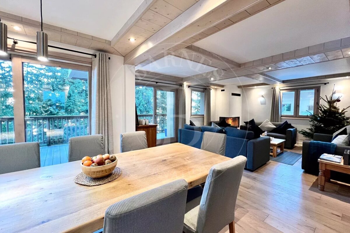 Cozy open-plan living and dining room with a large wooden table, blue sofas, a lit fireplace, and a Christmas tree, overlooking a forest.