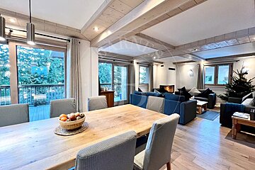 Cozy open-plan living and dining room with a large wooden table, blue sofas, a lit fireplace, and a Christmas tree, overlooking a forest.