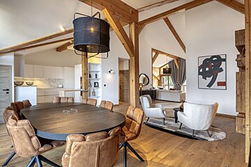 A chic, open-plan interior showing a dining room with a large table, a kitchen, and a living area with white armchairs, featuring exposed wooden beams.