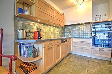 A cozy kitchen featuring light wood cabinets, green tiled backsplash and floor, equipped with modern appliances, and shelves with books and games.