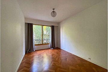 An empty room with herringbone patterned wooden floors, white walls, and a large window looking out onto green trees.