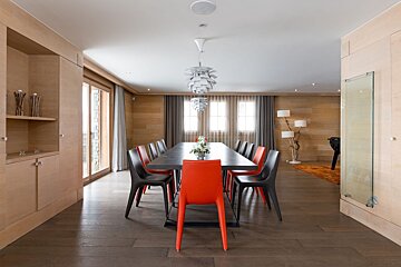 Modern dining room with a long dark table, alternating black and orange chairs, and distinctive pendant lights. Wood paneling and large windows create a warm ambiance.
