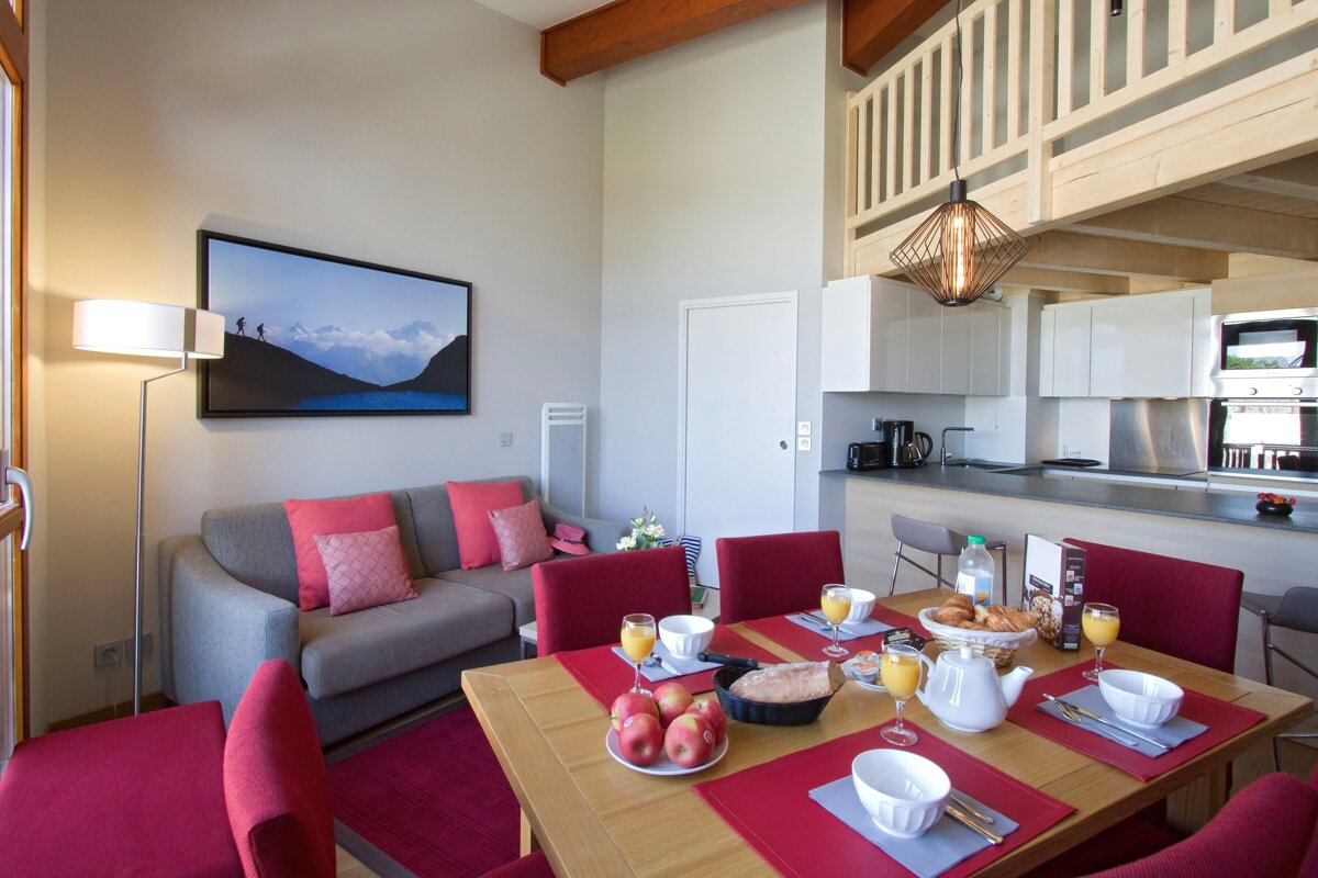A modern, open-concept living space with a dining area set for breakfast, a gray sofa with red cushions, and a white kitchen, featuring a loft above.