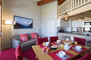 A modern, open-concept living space with a dining area set for breakfast, a gray sofa with red cushions, and a white kitchen, featuring a loft above.