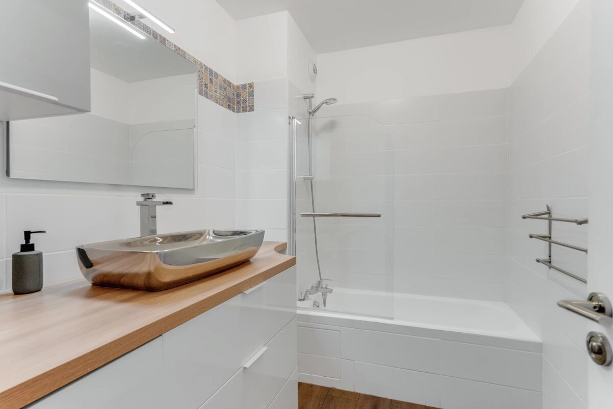 Modern white bathroom featuring a metallic vessel sink on a wooden vanity and a bathtub with a glass screen.