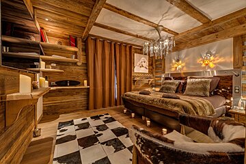 A cozy, rustic wooden bedroom featuring a bed with fur throws, a crystal chandelier, and a checkered cowhide rug.