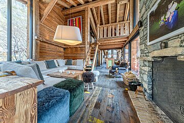 Cozy rustic chalet interior featuring wooden beams, a stone fireplace, fuzzy ottomans, a large sectional sofa, and a loft staircase.