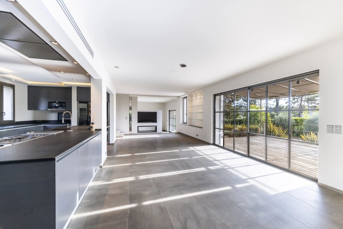 Modern, open-plan home interior with a sleek dark kitchen, tiled floors, and large glass sliding doors opening to a sunny outdoor deck.