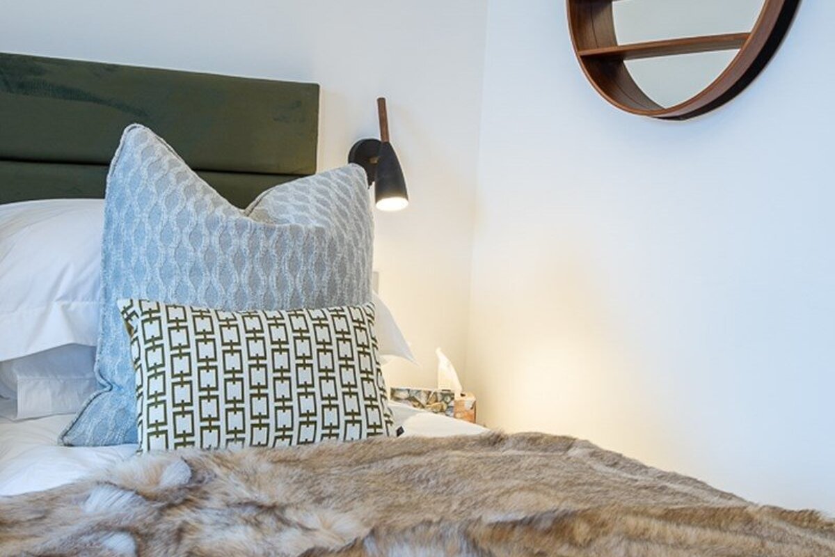 A bed styled with textured pillows, a faux fur throw, a green headboard, a reading lamp, and a round wall mirror.