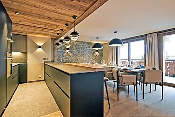 Chic open-plan kitchen and dining space featuring a wooden ceiling, stone accent wall, modern lighting, and large windows.