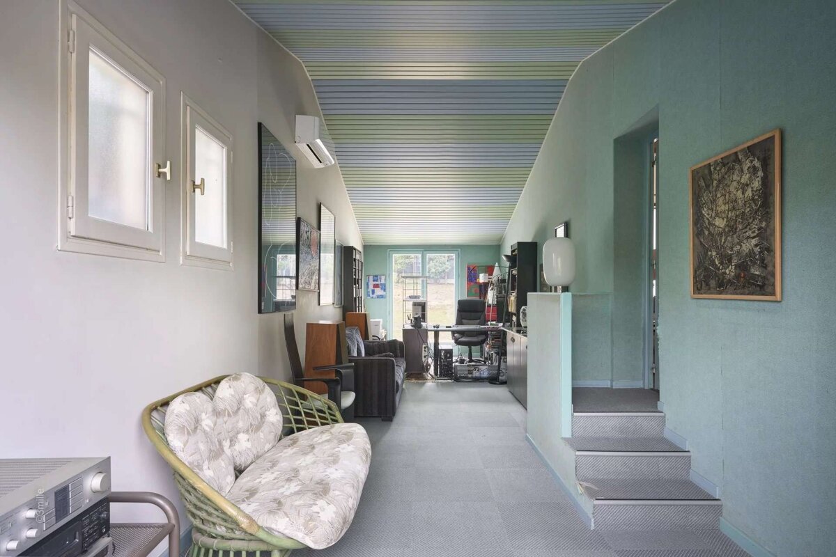 A long, narrow room with light blue/green walls, striped ceiling, grey floor, rattan sofa, office area at the end, and steps.