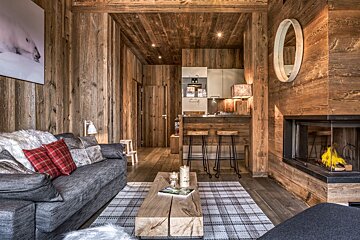 A warm, rustic living space with wood paneling, a gray sofa, plaid rug, cozy fireplace, and a small kitchen bar.