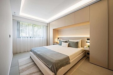 A serene, modern bedroom with neutral tones. It has an integrated bed frame/headboard, recessed and ambient lighting, and sheer curtains over a window.