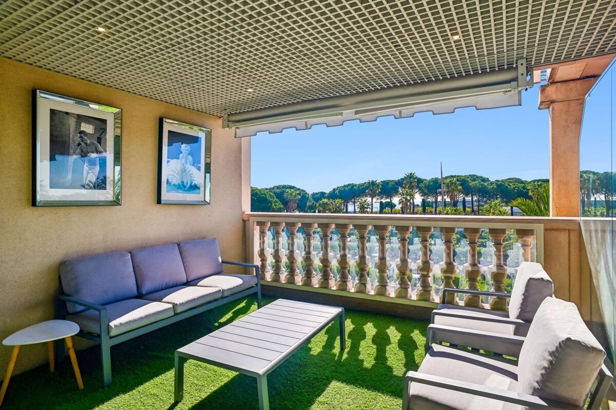 A sunny balcony with outdoor seating on artificial grass, framed art, and a balustrade, overlooking trees against a clear blue sky.