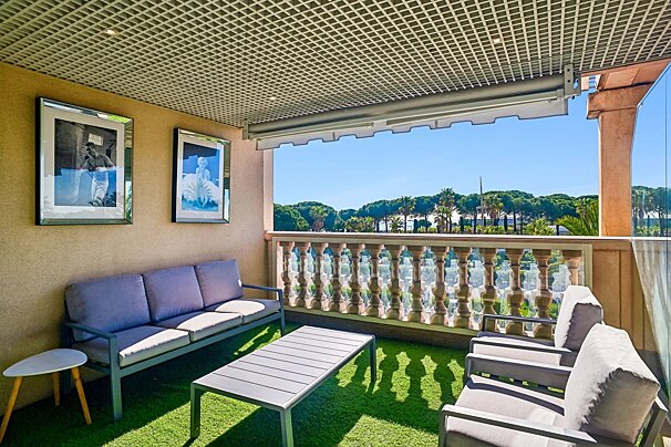 A sunny balcony featuring grey outdoor furniture on artificial grass, framed wall art, and a balustrade overlooking trees.