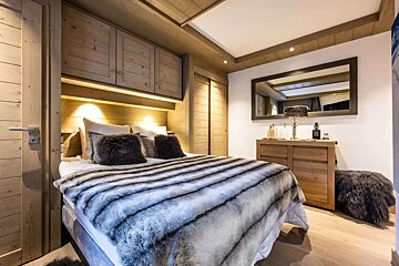 A chic and cozy bedroom with rustic wood paneling, a large bed covered in a striped furry blanket, built-in storage, and a dresser with a mirror.