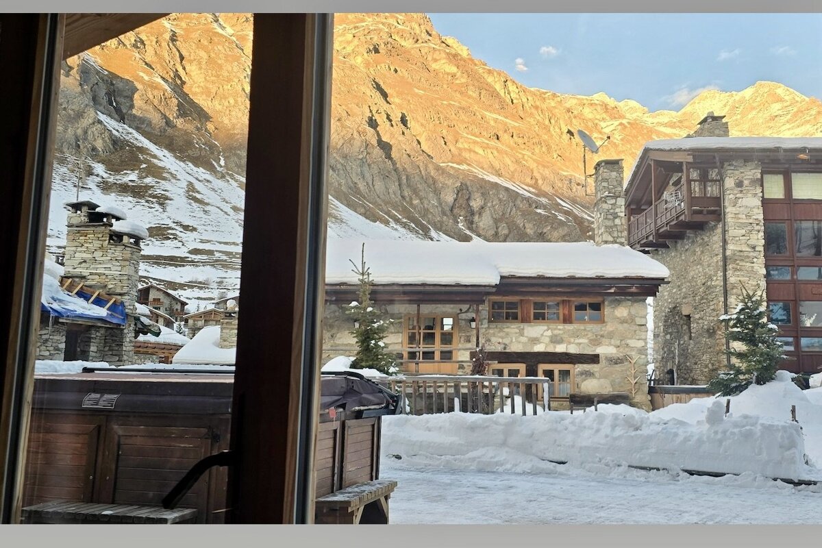 A view through a window of snow-covered stone chalets in an alpine village under towering, sunlit mountains.