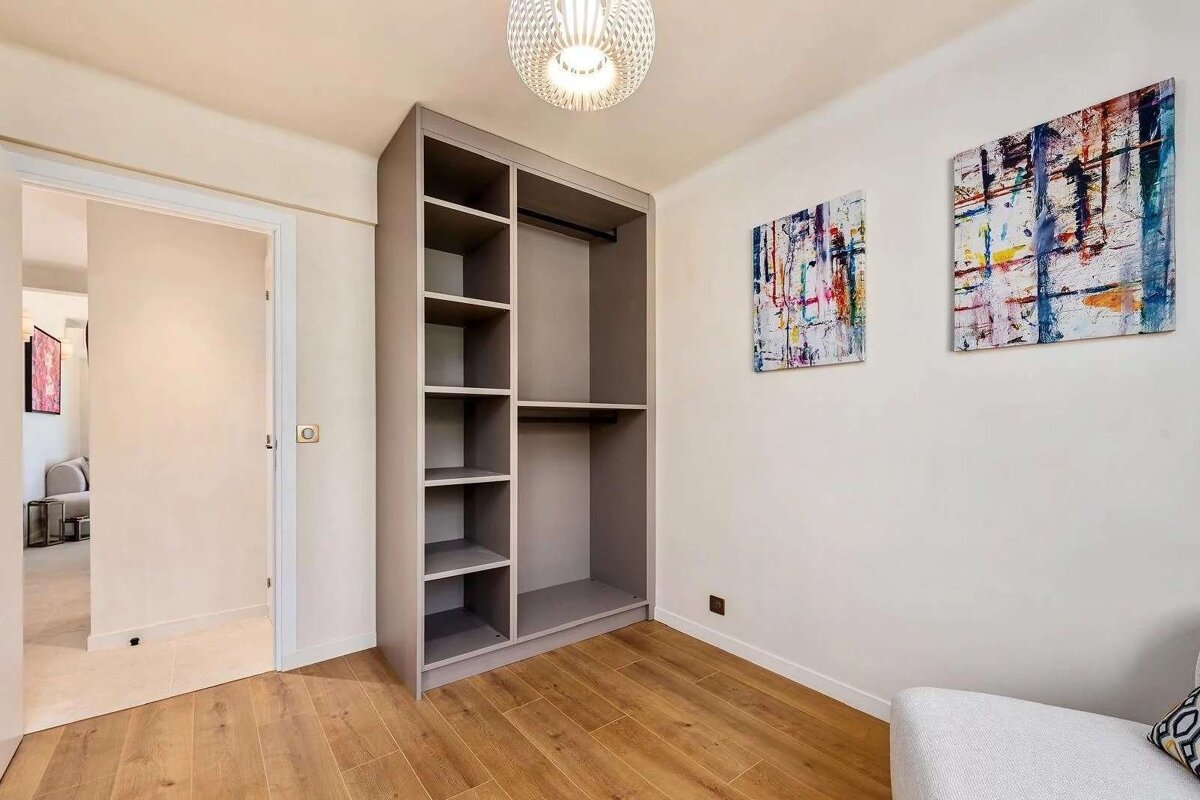 A modern room with light wood floors, a grey built-in open wardrobe, and two colorful abstract paintings on the white wall. A stylish ceiling light hangs above.