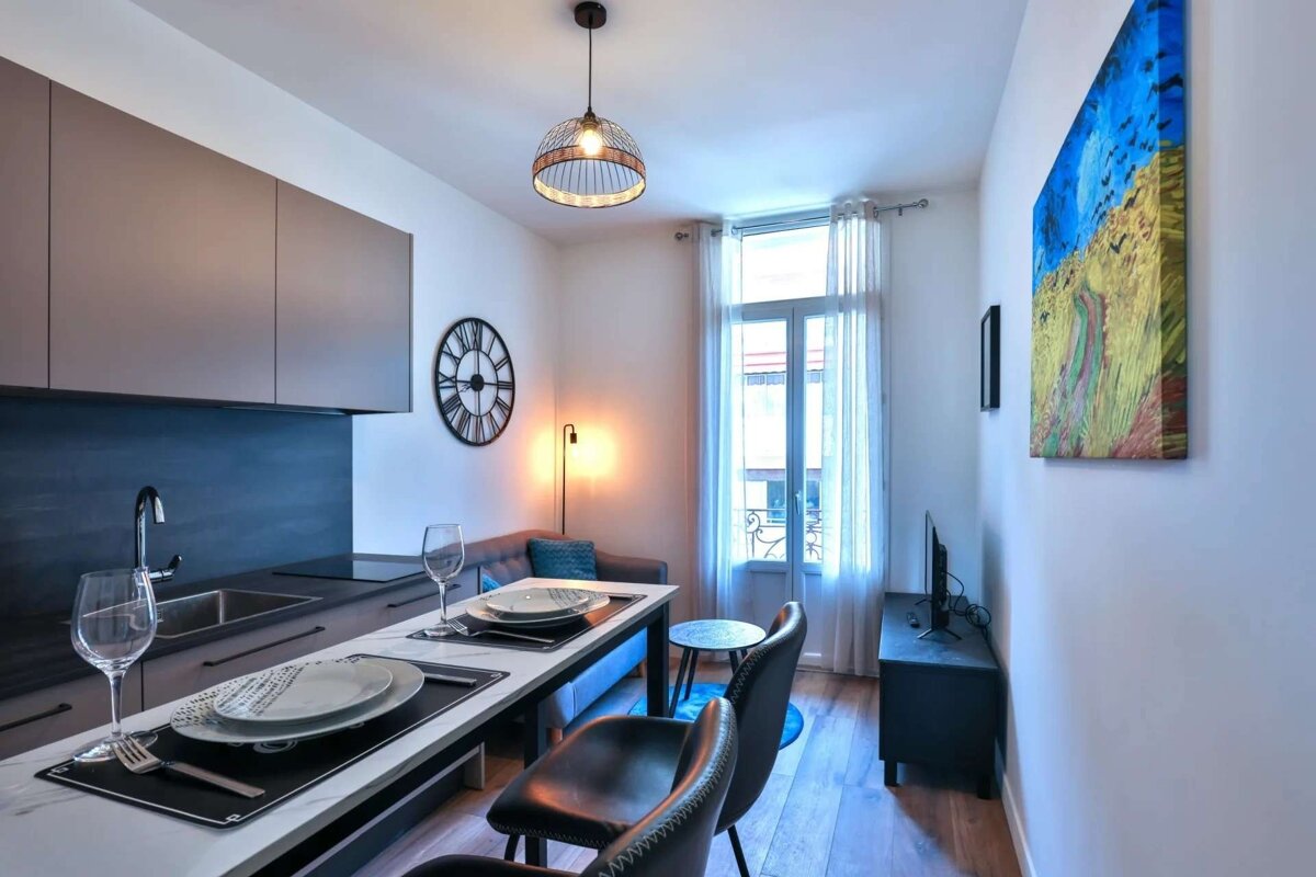 A modern, compact studio apartment featuring a sleek kitchen, dining table, sofa, large window, and colorful abstract artwork.