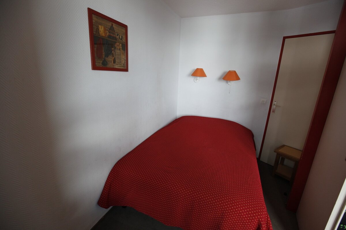 A simple bedroom with white walls, a red-covered single bed, two orange wall lights, a picture, and a door with red trim.