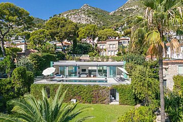 A sleek modern glass house with a poolside terrace, surrounded by lush greenery and palm trees, nestled against a mountainous backdrop.