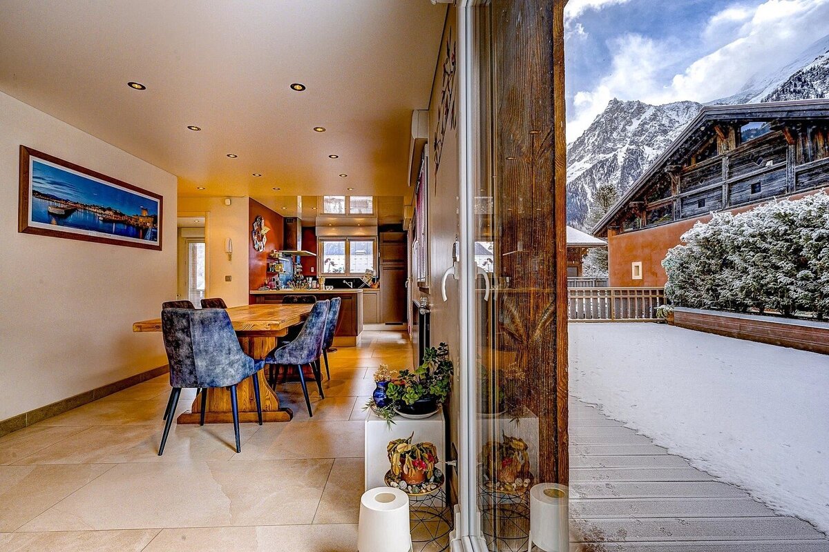 Bright indoor dining/kitchen area connected to a snowy outdoor scene with mountains and an alpine chalet through a large glass door.