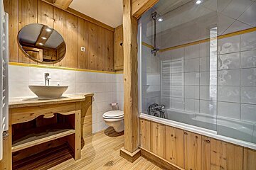 A rustic, wood-paneled bathroom with white tiles, yellow trim, a bowl sink, toilet, and a bathtub with a glass screen.