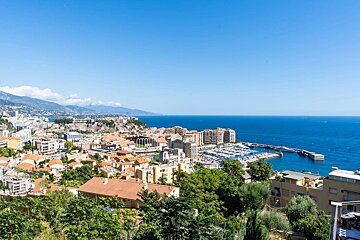 A sunny high-angle view of a coastal city with a marina, red-roofed buildings, lush trees, and a deep blue sea under a clear sky.