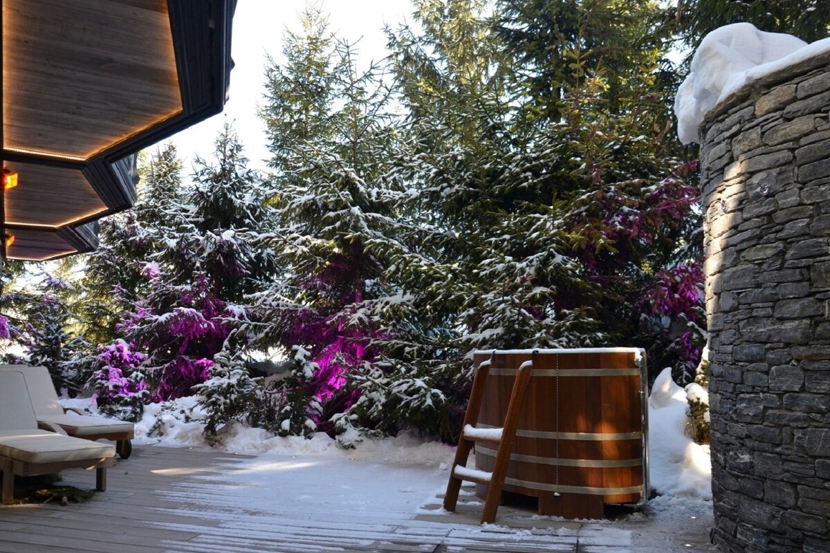 A wooden hot tub is surrounded by snow covered trees