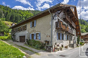 Rustic B&B house with wooden balconies & blue shutters, set against a backdrop of dense green mountains & a bright blue sky.