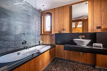 A modern rustic bathroom featuring warm wood paneling, sleek grey marble tiles, a white bathtub, vessel sink on a black counter, and a pebble floor.