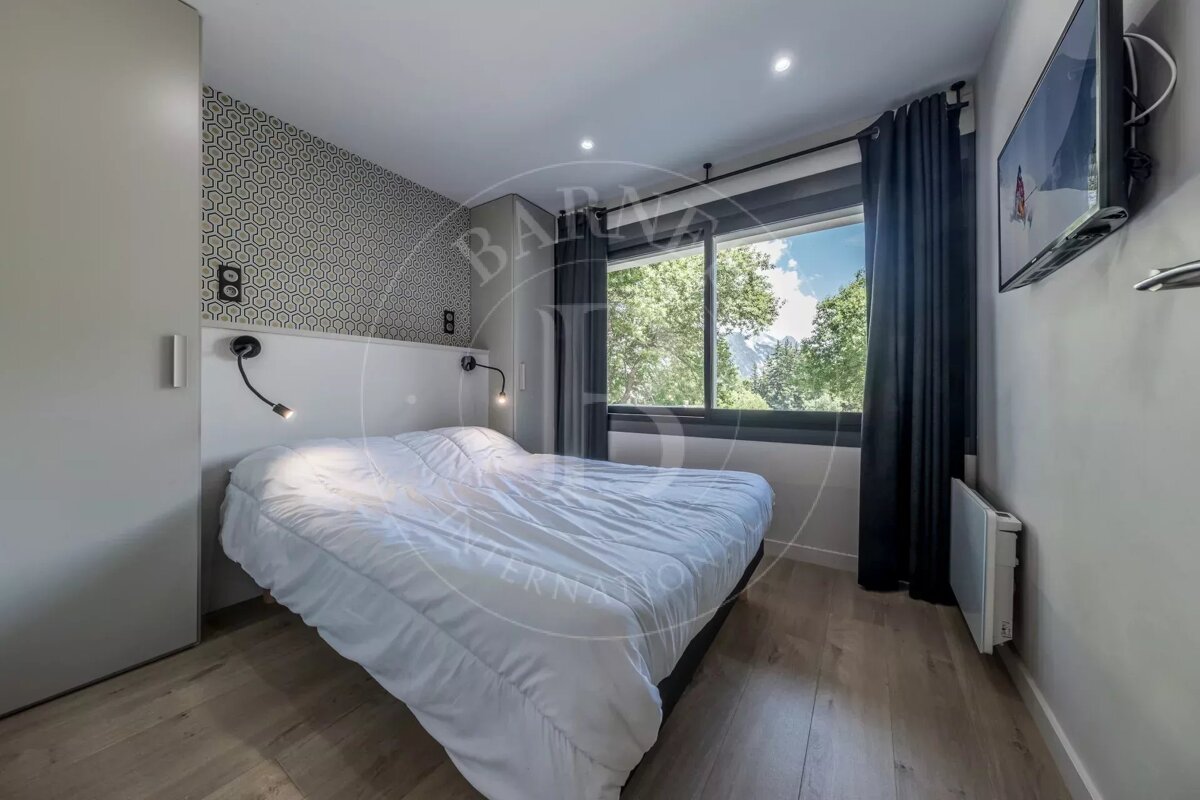 A modern bedroom with a white bed, large window showcasing a mountain and forest view, wall-mounted TV, wooden floor, and geometric wallpaper.
