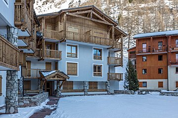 Snow-covered alpine resort buildings with wooden balconies, stone foundations, and light facades, set against a mountainous backdrop.