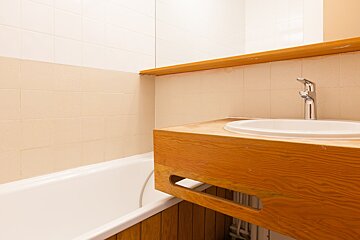 A bathroom features a white bathtub on the left and a wooden vanity with an oval sink and chrome faucet on the right. Tiled walls complete the scene.