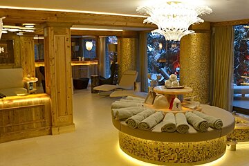A cozy, luxurious spa lounge featuring warm wood, gold mosaic accents, soft lighting, rolled towels on a round bench, recliners, and large windows to a snowy landscape.
