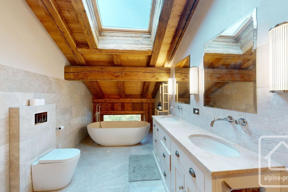 A chic bathroom with a freestanding tub, double vanity, and toilet, featuring rustic wooden beams, a skylight, and light tiles.