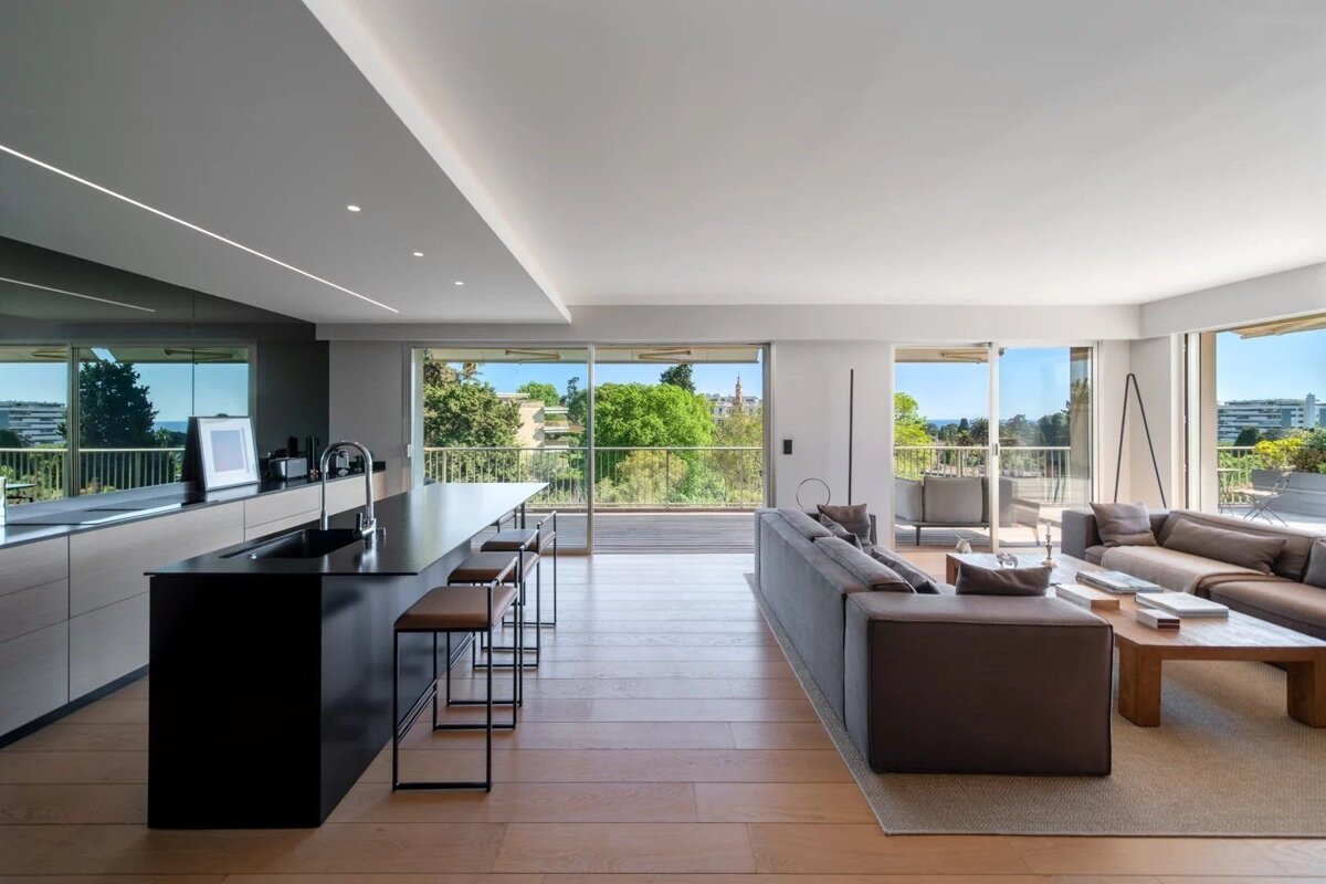 A modern open-plan living room and kitchen featuring large windows, wood floors, contemporary furniture, and a balcony with lush green views.