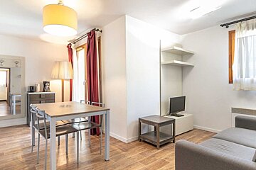 Bright apartment interior featuring a dining table, a grey sofa, a small TV, and wooden flooring.