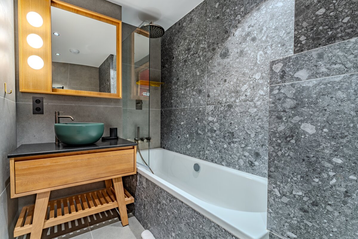 Modern bathroom with gray speckled tiles, wooden vanity, green vessel sink, and a bathtub with shower.