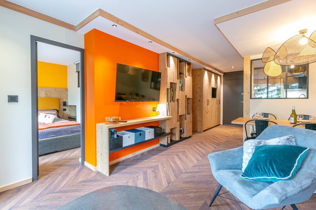 Warm, modern living space featuring a bright orange wall with TV, intricate wooden shelving, herringbone floor, and a view into a cozy bedroom.
