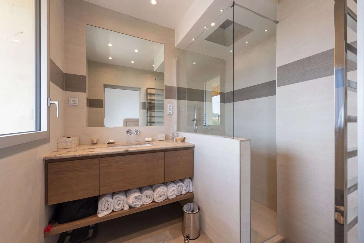 Modern bathroom with a glass walk-in shower, wood vanity, large mirror, and light-colored tiles with a grey stripe. Clean and contemporary.