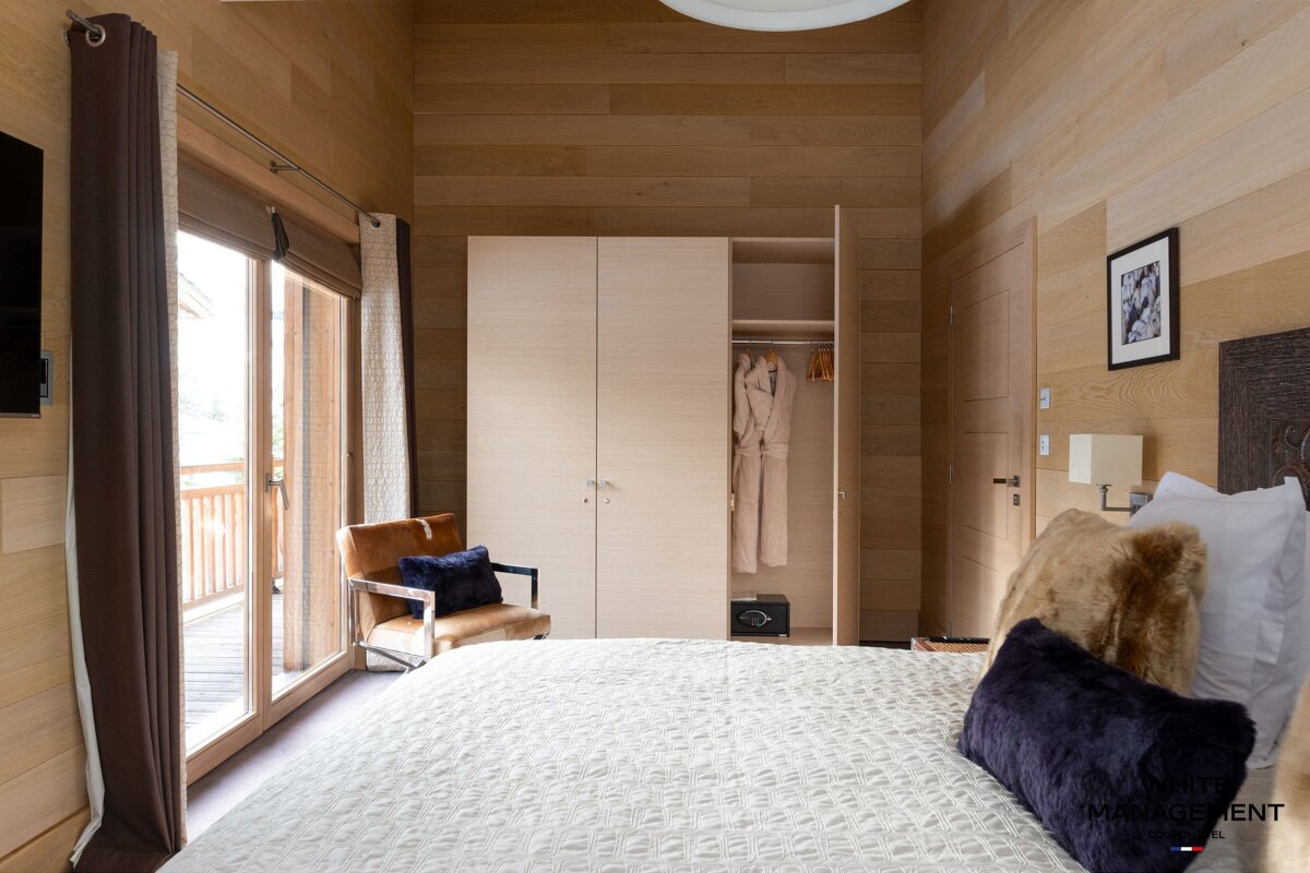 A cozy, modern bedroom with wood-paneled walls, a large bed, an open wardrobe with bathrobes, an armchair, and a balcony view.